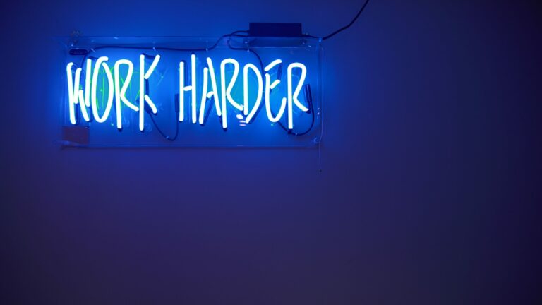 Blue neon off-set at the top right of a blue wall. Text reads, "Work harder."
